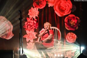 Scena. Kobieta w wieku seioralnym siedzi na białym, wiklinowym fotelu. Ubrana jest w czerwoną sukienkę. W prawej ręce trzyma mikrofon, do którego śpiewa. W tle dekoracja z dużych białych i ćzerwonych kwiatów z papieru, tworzących girlandę. Na dole sceny jasne światło reflektorów. 