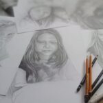 Kartki papieru z rysunkami, w centrum portret dziewczynki. Poprawej strinie położonych kilka ołówków