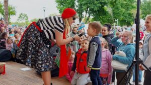 Kobieta przebrana w stój piratki nachyla się nad małym chłopcem. Trzyma w ręce mikrofon, do którego chłopiec coś mówi. W tle grupa innych dzieci.