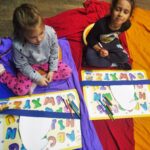 Dwie kulkuletnie dziewczynki siedzą na rozłożonej na podłodze kolorowej chuście animacyjnej. Pzed nimi leżą tabliczki z alfabetem.