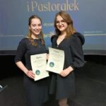 Dwie dziewczynki stoją bokiem do siebie. W rękach trzymają dyplomy. Z tyłu napis Festiwal Kolęd i Pastorałek.