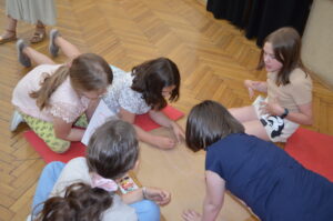 Kilkoro dzieci pochyla się nad rozłożoną na podłodze dużą kartką paieru. Coś na niej rysują.