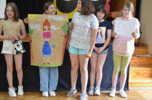 Kilka dziewczynek stoi obok sieboie. Jedna trzyma dużą kartkę papieru z rysunkiem postaci.