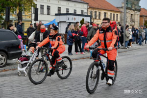 Kobieta i mężczyzna jadący obok siebie na rowerach. Ubrani są w jednakowe uniformy w kolorze pomarańczowym.