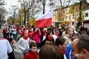 Grupa osób w różnym wieku biegnąca po ulicy miasta. W centrlanym punkcie zdjęcia flaga biało-czerwona.