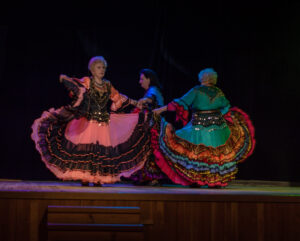 Trzy starsze kobiety tańczą na scenie. Na sobie mają bogato zdobione cygańskie suknie. Tańczą razem, trzymając się za ręce. Na ich twarzach widać skupienie.