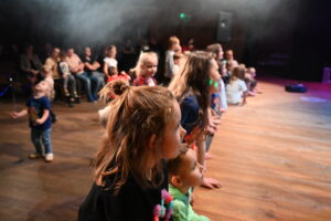 Dzieci stojące przy scenie z uwagą patrzą w stronę sceny. W tle za nimi rodzice siedzą na widowni.