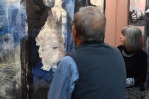 Starszy mężczyzna i kobieta stoją przed obrazem, który wisi na ścianie przed nimi. Ze skupieniem oglądają wystawioną pracę.