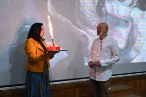 Kobieta i starszy mężczyzna stoją na przeciwko siebie. Kobieta trzyma w rękach tort, na którym odpalona jest raca. Mężczyzna stoi na przeciwko i uśmiecha się.
