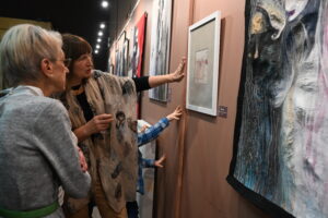 Dwie starsze kobiety przyglądają się obrazom z tkaniny artystycznej. Obrazy wiszą na ścianie przed nimi. Jedna z kobiet wyciąga dłoń w kierunku pracy, wskazując na nią. Kobiety wspólnie interpretują sztukę, na ich twarzach widać zainteresowanie i skupienie. 