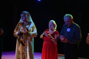 Trzy starsze osoby stoją na scenie. W rękach trzymają jabłko, które obierają małym nożykiem. Na ich twarzach widać skupienie. 