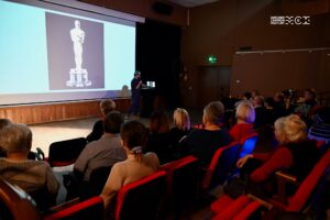 Sala kinowa. Na widowni widać kilka osób. Przed nimi duży jasny ekran, a na nim wyswietlony obraz statuetki Oscara.