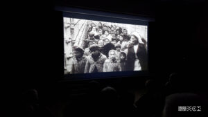Ekran kinowy. Na nim wyświetlony kadr z filmu przedstawiający więźnió∑ obozu koncentracyjnego. Niektórzy są w pasiakach, inni w cywilnych ubraniach.