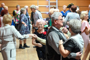 Grupa seniorów, kobiet i mężczyzn znajduje się w dużej sali. Osoby na zdjęciu podobierane są w pary i wykonują ruch przypominający taniec. Wszyscy szeroko uśmiechają się do siebie, na ich twarzach widać dobrą zabawę.