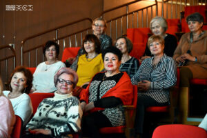 Grupa kobiet w senioralnym wieku. Siedzą na krzesłach ustawionych jak na widowni w teatrze. Kobiety uśmiechają się i patrzą przed siebie.