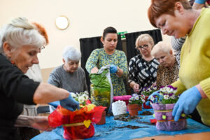 Grupa seniorek stoi wokół stołu, na którym stoją kolorowe donice z kwiatami oraz worki z ziemią. Wydaje się, że kobiety przesadzają rośliny. Pomaga im w tym dorosła kobieta ubrana w rękawice. Na twarzach uczestników widać pełne skupienie.