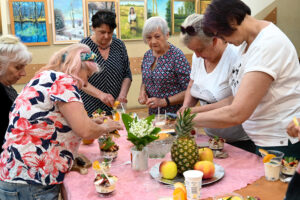 Kobiety w wieku senioralnym stoją wokół stolika, na którym znajdują się owoce i jednorazowa zastawa. Wyglądają, jakby przygotowywały jakiś posiłek. Na ich twarzach widać skupienie i zadowolenie. 