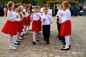 Dzieci ubrane w stroje w barwach narodowych. Część z nich tworzy szpaler. środkiem idą dziewczynka i chłopiec, trzymają się za wyciągnięte przed siebie ręce. Dziewczynki mają w rękach czerwone róże.