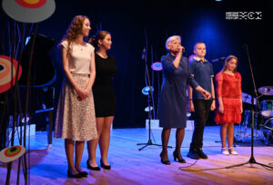 Grupa osób stojąca na scenie. Są w różnym wieku i różnej płci. Najstarsza kobieta śpiewa lub mówi do mikrofonu.