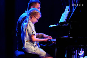 Dziewczynka i młody mężczyzna przy fortepianie. Grają na cztery ręce.