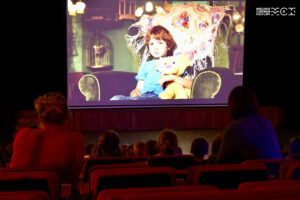 Sala kinowa, w tle ekran z wyświetlanym na nim filmem. 
