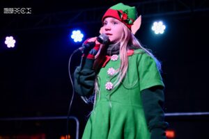 Dziewczynka w stroju elfa. W ręce trzyma mikrofon, śpiewa lub mówi.