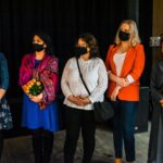 Pięć kobiet, stoją obok siebie. Mają maseczki higieniczne na twarzach. Jedna trzyma bukiet kwiatów.