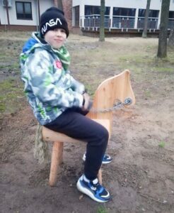 Kilkuletni chłopiec siedzi na drewnianym koniu. Ubrany jest w kolorową kurtkę. Na głowie ma czapkę. Patrzy w prawo.