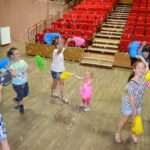 Kilkoro dzieci tańczy z kolorowymi pomponami na sali widdowiskowej.