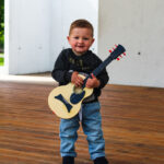 Mały chłopczy z gitarą w ręce. Paatrzy na wprost, lekko się uśmiecha.