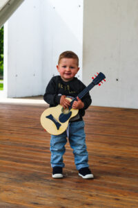 Mały chłopczy z gitarą w ręce. Paatrzy na wprost, lekko się uśmiecha.