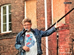 Mężczyzna w średnim wieku. Nosi okulary. Stoi przy mikrofonie, do którego coćś mówi. Ręką wskazuje na mur starego budynku czerwonej cegły.