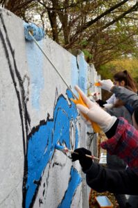 Fragment betonowego ogrodzenia. Grupa młodych ludzi maluje na nim mural.