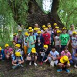 Grupa dzieci pozuje do zdjęcia. Na głowach mają żółte czapki z daszkiem. W tle drzewa okryte zielonym liśćmi.