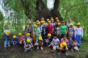 Grupa dzieci pozuje do zdjęcia. Na głowach mają żółte czapki z daszkiem. W tle drzewa okryte zielonym liśćmi.