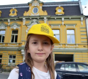 Dziewczynka w żółtej czapce z daszkiem. W tle zabytkowa kamienica