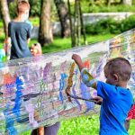Kiluletni chłopiec maluje farbami na folii stretch rozciągniętej między pnami drzew w parku.