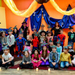 Kilkudiesięcioosobowa grupa dzieci w przebraniach czarownic, wróżek i wróżbitów. Częśc z nich stoi, część siedzi na dywanie. W tle kolorowa dekoracja.