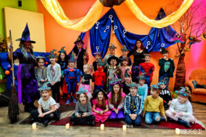 Kilkudiesięcioosobowa grupa dzieci w przebraniach czarownic, wróżek i wróżbitów. Częśc z nich stoi, część siedzi na dywanie. W tle kolorowa dekoracja. 