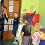 Przed drzwiami stoi kobieta przebrana za czarownicę. Przed nią pięcioro dzieci w przebraniach andrzejkowych. Kobieta trzyma w ręce miotłę.