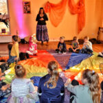 Grupa dzieci siedzi w kręgu na kolorowej chuście animacyjnej . Patrzą na dwie kobiety przebrane za czarownice.