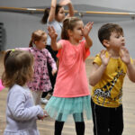 Grupa dzieci na sali tanecznej. Unoszą ręce do góry, nasladują ruchy pokazywane przez instruktora, przybierając pozy taneczne.