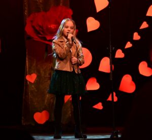 Dziewczynka stoi na scenie z mikrofonem w ręce. Śpiewa. Patrzy przed siebie. W tle scenografia z kwiatów i czerwonych serc.