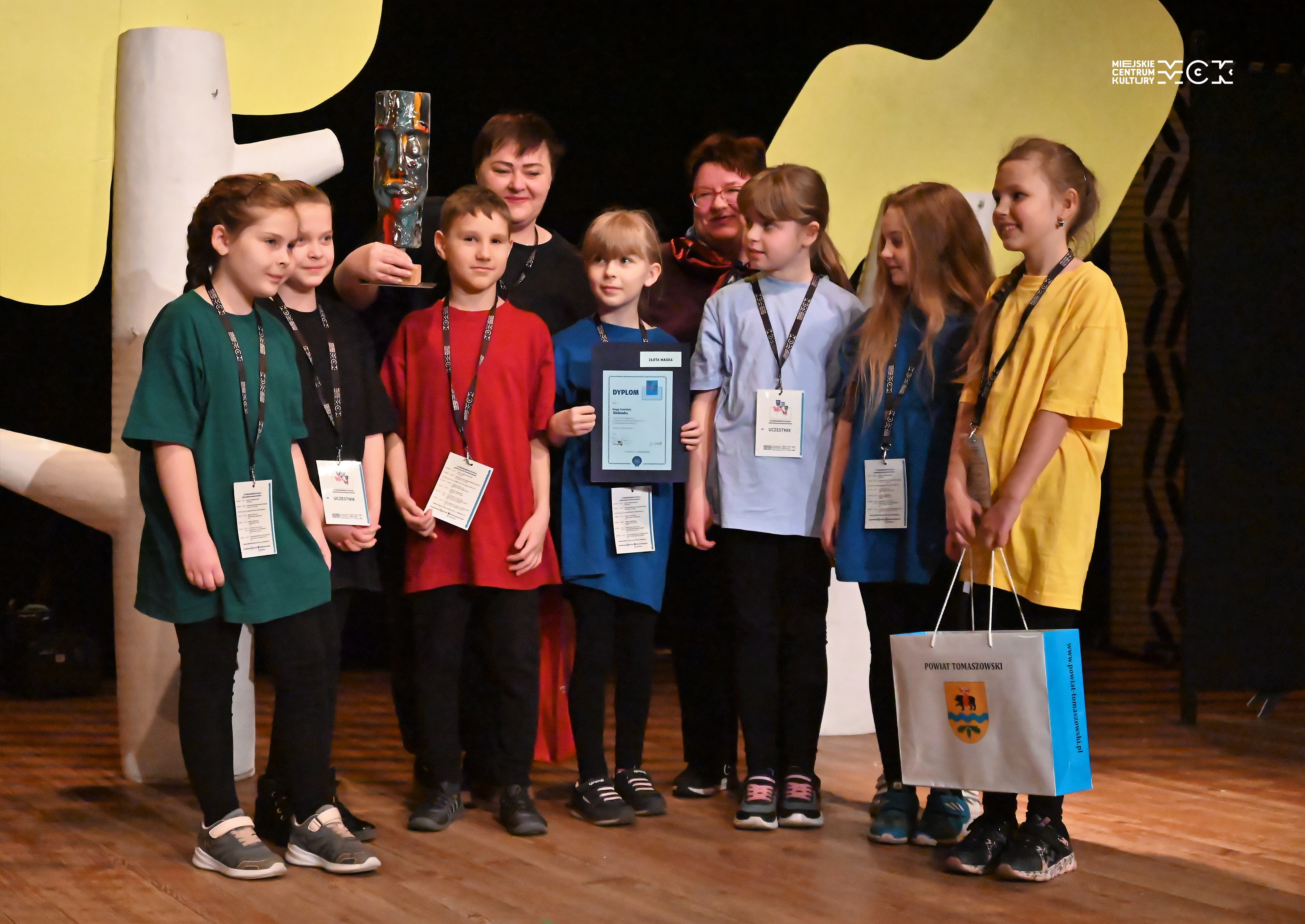 Grupa dzieci w kolorowych koszuklach stoi na scenie. Na szyjach zawieszone mają identyfikatory . Kobieta stojąca za nimi trzyma w ręce statutkę - maskę.