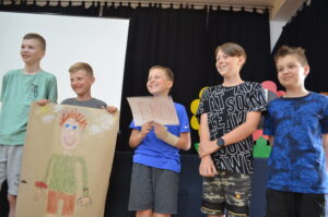 Czterech chłopców stoi obok siebie. Śmieją się. Dwóch trzyma przed sobą kartki papieru z rysunkiem postaci i napisem lans.