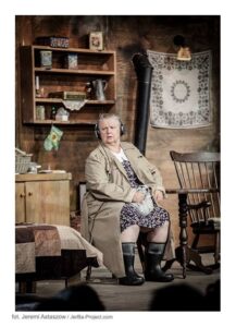 Starsza kobieta siedzi na krześle. Ubrana jest w jasny prochowiec i gumowe buty. Na uszach ma słuchawki. W tle za nią na ścianie wisi drewniamna półka z różnymi naczyniami.