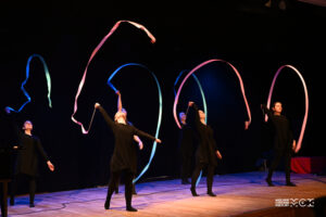 Na scenie znajdują się cztery młode osoby ubrane na czarno. W rękach mają kolorowe szarfy, którymi żonglują nad głowami. Scena oświetlona jest kolorowymi światłami.