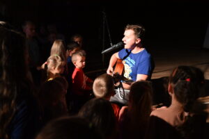 Dorosły mężczyzna siedzi na krawędzi sceny. Gra na gitarze oraz śpiewa do mikrofonu, który ustawiony jest przed nim. Wokół mężczyzny przy scenie stoją dzieci, które są w niego wpatrzone i z uwagą przysłuchują się artyście.