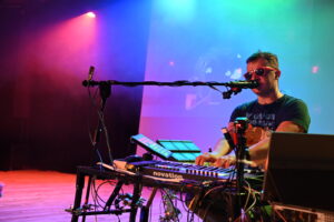 Mężczyzna siedzi przy keyboardzie, na którym gra. Na wysokości ust ma ustawiony mikrofon, do którego śpiewa. Scena oświetlona jest na kolorowo. Mężczyzna ma założone okulary przeciwsłoneczne.