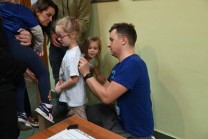 Dorosły mężczyzna podpisuje dziewczynce tył koszulki. Mężczyzna przykuca za dziewczynką i markerem składa autograf. Obok dziewczynki stoi dorosła kobieta oraz inna mała dziewczynka. Obie przyglądają się autografowi na koszulce.